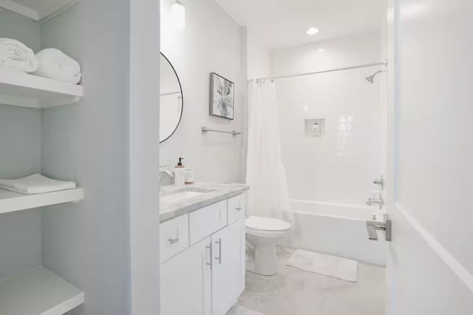 Bright modern white bathroom interior with marble-topped vanity and round mirror, open shelves with folded towels, toilet and tub-shower with white curtain