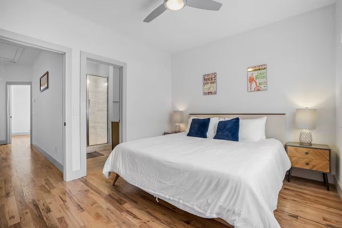 Bright modern master bedroom with king bed in crisp white linens, two navy throw pillows, wooden nightstands and lamps, hardwood floors, ceiling fan, and an open doorway to an ensuite shower.