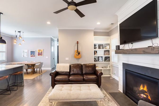 Modern open-concept living room with dark leather sofa, tufted ottoman, wall-mounted TV, cozy gas fireplace, acoustic guitar on the wall, and adjacent dining area