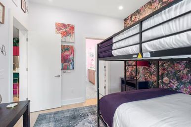 Bright modern bedroom with black metal bunk bed and purple bedding against floral wallpaper, colorful abstract art, area rug and open door to a pink-accented bathroom.