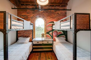 Cozy industrial-style bunk room with metal-and-wood bunk beds, two lower twin beds, exposed red brick wall with painted floral mural, arched window overlooking trees, wooden floor and central nightstand.