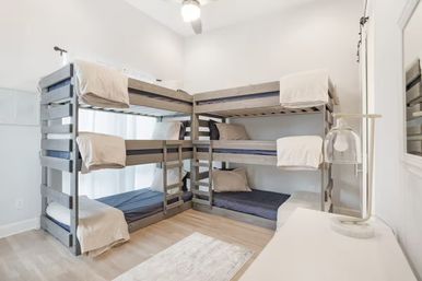 Bright airy bedroom with two gray L-shaped wooden bunk beds for six twin mattresses, neutral beige linens and navy sheets, light wood floors, ceiling fan, white dresser and bedside lamp.