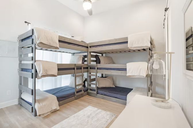 Bright airy bedroom with two gray L-shaped wooden bunk beds for six twin mattresses, neutral beige linens and navy sheets, light wood floors, ceiling fan, white dresser and bedside lamp.