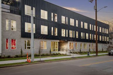 Dusk view of a sleek three-story modern apartment building with black metal cladding over a concrete base, warmly lit windows, sidewalk and urban street with utility poles and traffic cones.