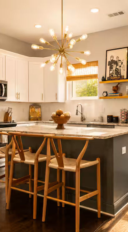 Sunlit modern kitchen with white cabinets, stainless steel fridge, marble-topped island and wooden breakfast bar stools under a starburst pendant light, hardwood floors and bamboo shades.
