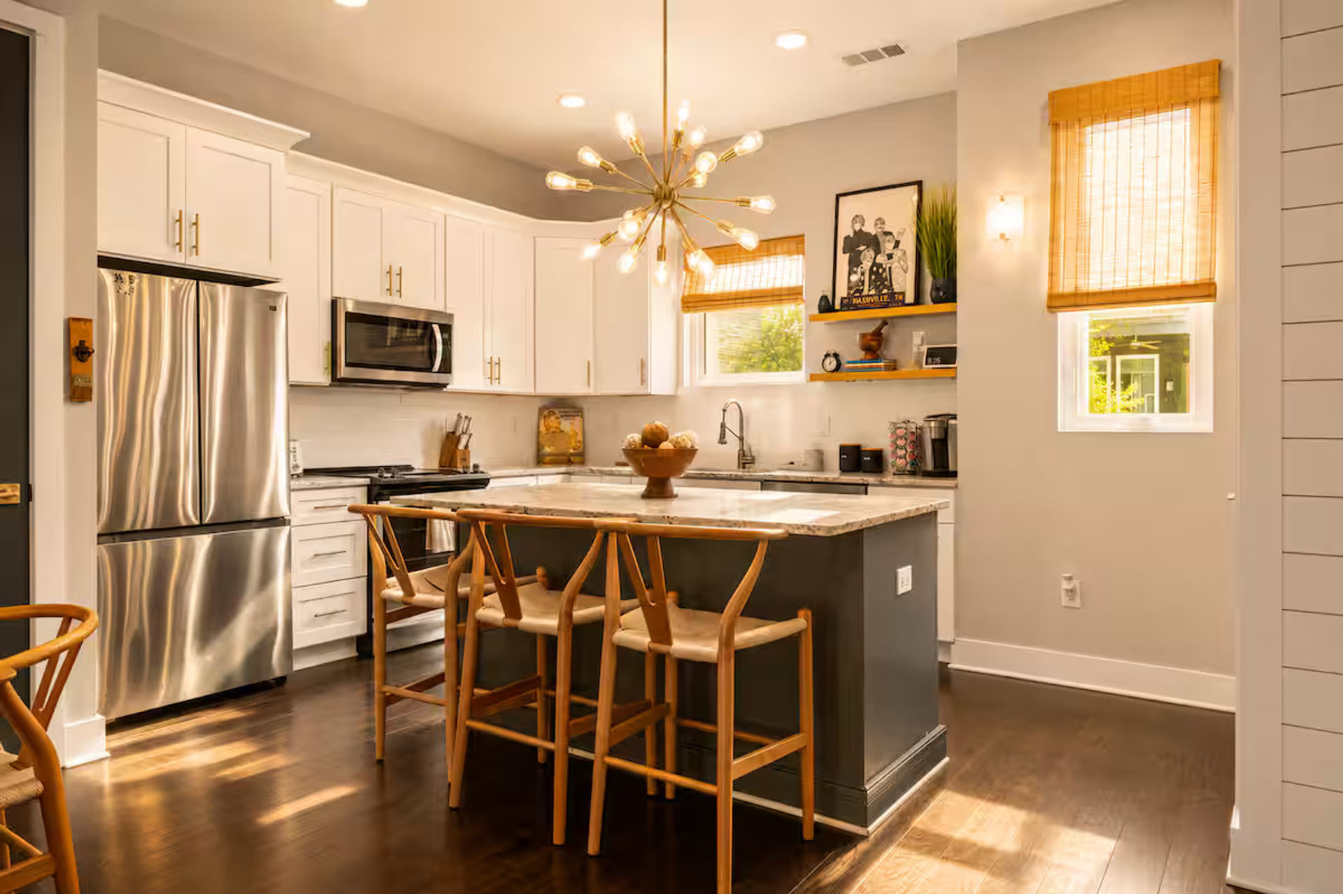 Sunlit modern kitchen with white cabinets, stainless steel fridge, marble-topped island and wooden breakfast bar stools under a starburst pendant light, hardwood floors and bamboo shades.