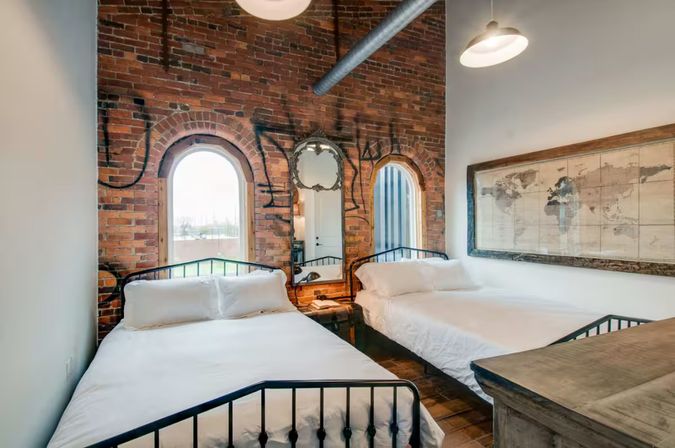 Bright industrial loft bedroom with exposed brick and arched windows, two metal-framed beds with white linens, an ornate mirror between windows and framed world map art