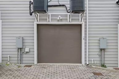Gray garage door set in light-gray horizontal siding with a paver driveway, two HVAC condensers mounted above and utility boxes flanking the entrance.