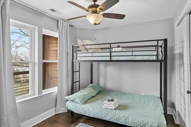 Cozy bright bedroom with black metal bunk bed, mint-green quilted bedding and folded towels on the lower bed, ceiling fan, hardwood floor and large windows with tree view.