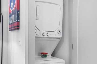 Cozy stacked white washer-dryer in a small laundry closet with a red-lidded container on top and colorful Nashville wall art on the nearby wall.