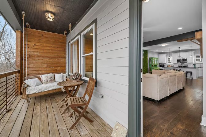 Cozy covered wooden balcony deck with rope-hung swing bench, round bistro table and folding chairs, opening into a bright open-plan living room and kitchen