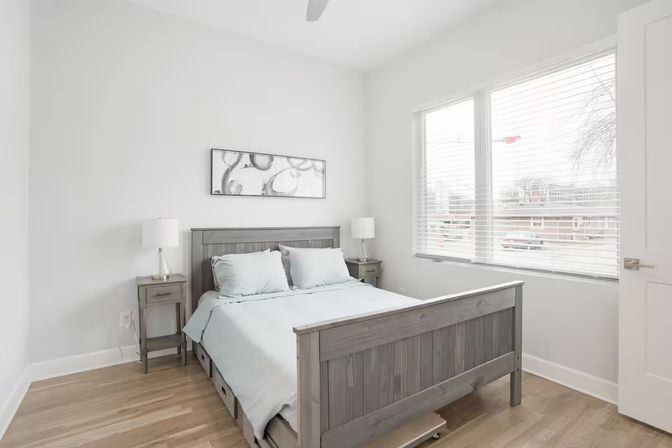 Bright, minimalist sunlit bedroom with gray wooden bed and matching nightstands, light-blue bedding, hardwood floors, large window with white blinds and modern table lamps.