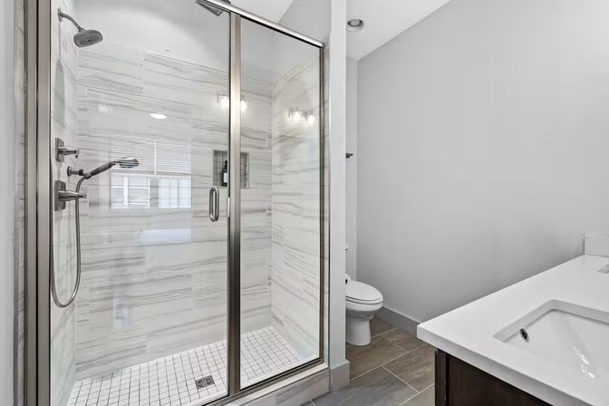 Contemporary gray-and-white bathroom with glass-enclosed tiled walk-in shower featuring rain and handheld showerheads, white rectangular sink vanity, toilet, and large porcelain floor tiles.