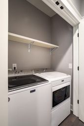 Compact laundry nook with white washer and matching dryer beneath a simple wood shelf, gray walls, white trim and hardwood floor.