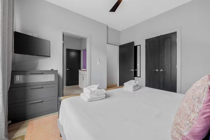 Contemporary urban bedroom with white linens and stacked towels on the bed, wall-mounted TV above a gray dresser, dark closet doors and an open doorway to the bathroom, pink accent pillow