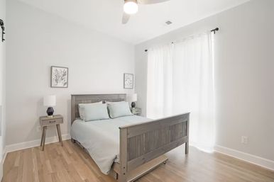 Sunny minimalist bedroom interior with gray wooden bed, pale blue bedding, matching nightstands and lamps, sheer white curtains and light hardwood floors