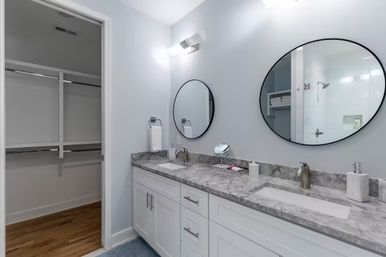 Bright contemporary master bathroom with marble double-sink vanity, white shaker cabinets, two round black-framed mirrors, chrome faucets and an open walk-in closet — sleek, spa-like design.