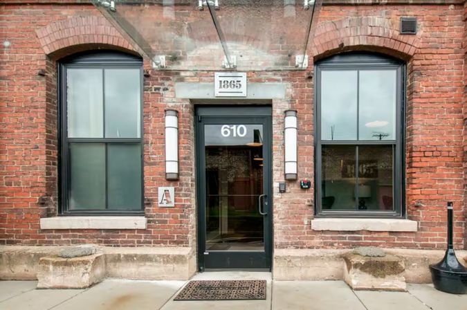 Inviting glass entrance numbered 610 in a restored 1865 red-brick building, flanked by tall windows and modern wall sconces on a city sidewalk.