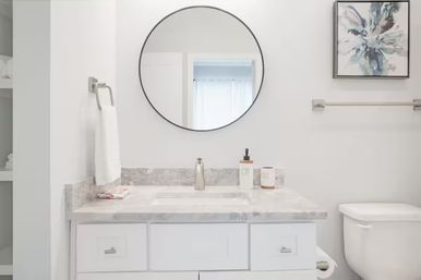 Bright modern white bathroom with marble-look vanity, chrome faucet, round wall mirror reflecting a window, towel on a ring, soap dispenser and abstract wall art beside a white toilet.