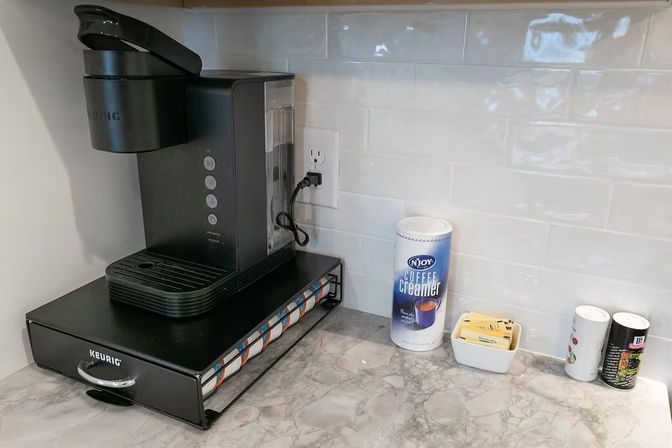 Compact kitchen coffee station with a black single-serve pod coffee maker on a K-cup drawer organizer, marble countertop and white subway tile backsplash; creamer canister, sugar packets and spice shakers to the right.