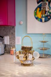 Cheery gold-rimmed porcelain tea cups and saucers on a tiered gold stand on a glossy kitchen countertop, with pastel blue wall, pink cabinet and colorful guitar wall art in the background.
