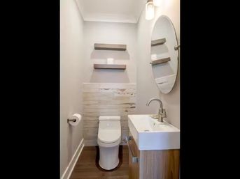 Cozy modern powder room interior with compact white toilet, rectangular wall sink on a wood vanity, round mirror, two floating wood shelves, shiplap wainscoting, single sconce light, and dark hardwood floor.