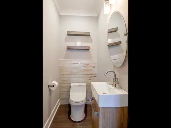 Cozy modern powder room interior with compact white toilet, rectangular wall sink on a wood vanity, round mirror, two floating wood shelves, shiplap wainscoting, single sconce light, and dark hardwood floor.