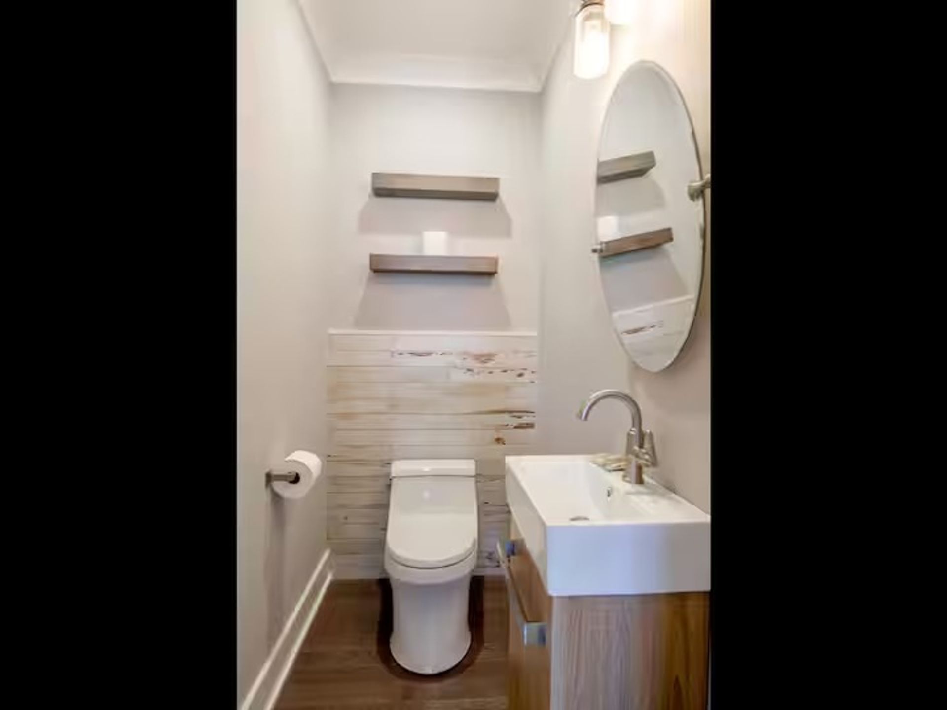 Cozy modern powder room interior with compact white toilet, rectangular wall sink on a wood vanity, round mirror, two floating wood shelves, shiplap wainscoting, single sconce light, and dark hardwood floor.
