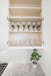 Open white kitchen cabinet in a modern kitchen with neatly arranged wine glasses and tumblers on shelves, marble countertop, white subway tile backsplash and small potted green plant.