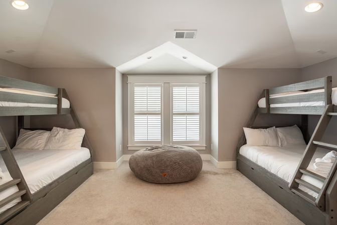 Family-friendly finished-attic bunk bedroom with matching gray wooden bunk beds, white linens, a large round gray beanbag in the center, beige carpet, and a window with white plantation shutters.