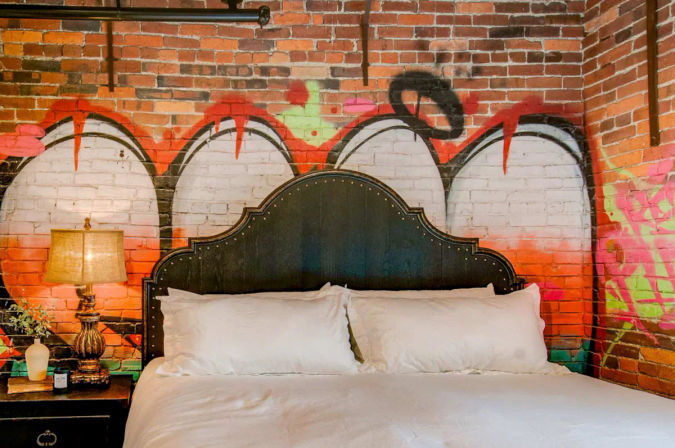 Urban loft bedroom with exposed red brick and playful graffiti mural behind a dark studded headboard, crisp white bedding and a vintage lamp on the nightstand.