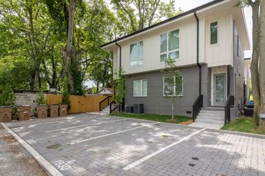 Modern two-story townhouse with gray brick and white siding, front steps and glass door, reserved brick-paver parking spots, row of curbside trash bins, wooden fence and large shade trees in a leafy suburban residential setting.