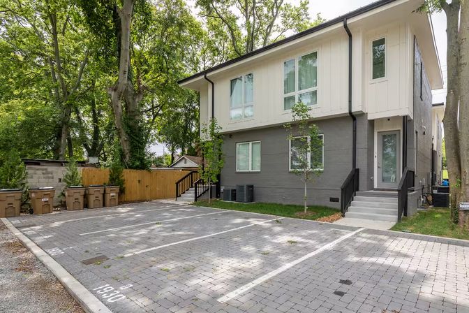 Modern two-story townhouse with gray brick and white siding, front steps and glass door, reserved brick-paver parking spots, row of curbside trash bins, wooden fence and large shade trees in a leafy suburban residential setting.
