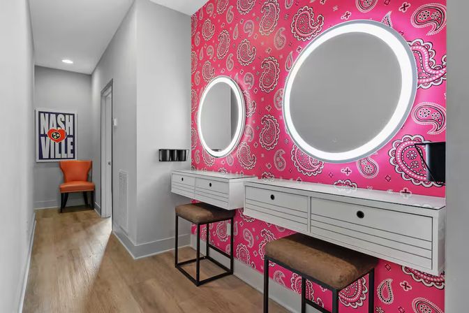 Vibrant dressing area with pink paisley wallpaper, two round LED-lit mirrors above floating white vanities, brown stools and wood floor, orange chair and 'Nashville' poster in the hallway.