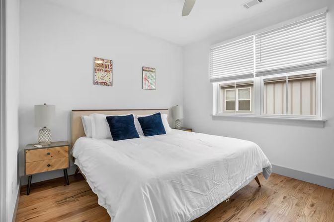 Sunlit modern minimalist bedroom with white walls and hardwood floors, large bed in white linens with two navy throw pillows, matching mid-century nightstands and lamps, and a wide window with white blinds.