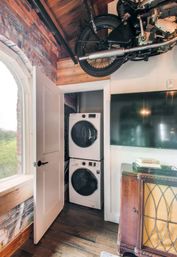 Loft-style living room with stacked washer and dryer tucked in a closet, exposed brick and wood ceiling, hanging vintage motorcycle, arched window and hardwood floors.