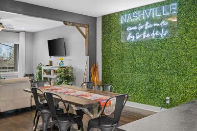 Modern Nashville apartment dining area with rustic wooden table, black metal chairs, open living room, TV, plants, and neon 'NASHVILLE' sign on a green moss wall.