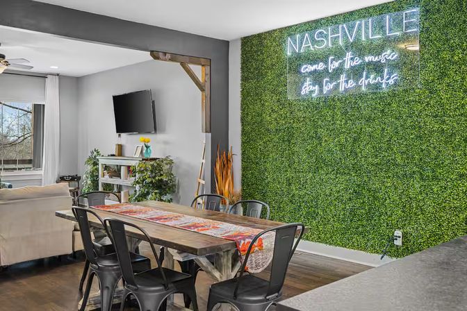 Modern Nashville apartment dining area with rustic wooden table, black metal chairs, open living room, TV, plants, and neon 'NASHVILLE' sign on a green moss wall.