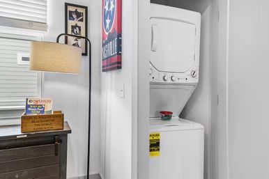Cozy laundry nook with a white stacked washer-dryer in a closet, a tall floor lamp, wooden crate of magazines on a dark console, and a Nashville poster on the wall.