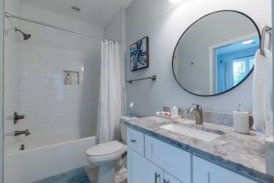 Bright modern residential bathroom with marble countertop vanity, round black-framed mirror, white subway-tiled bathtub with shower and curtain, toilet, blue bath rug and abstract blue wall art