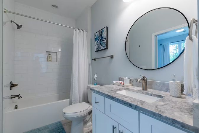 Bright modern residential bathroom with marble countertop vanity, round black-framed mirror, white subway-tiled bathtub with shower and curtain, toilet, blue bath rug and abstract blue wall art