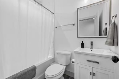Bright modern small bathroom interior with white tub and shower curtain, white toilet, white vanity sink with chrome faucet and mirror, gray hand towels and black soap dispenser