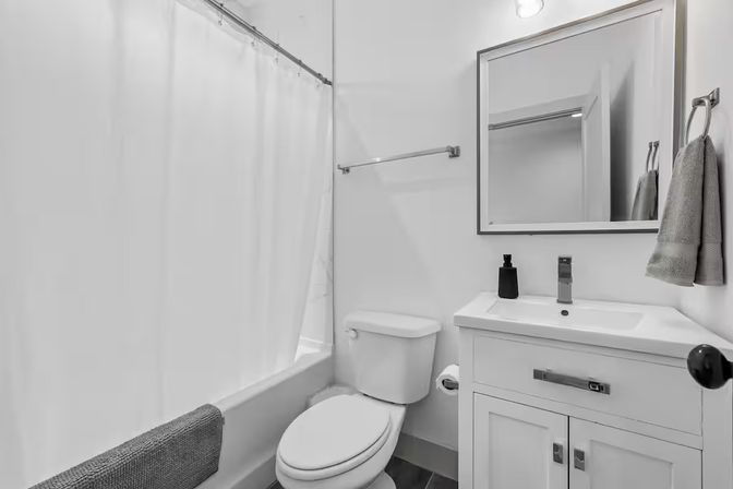 Bright modern small bathroom interior with white tub and shower curtain, white toilet, white vanity sink with chrome faucet and mirror, gray hand towels and black soap dispenser