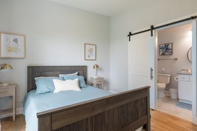 Cozy modern bedroom interior with dark wooden bed, light blue bedding and white accent pillow, matching nightstands with brass lamps, minimalist wall art, hardwood floors, and a white sliding barn door open to a tiled en‑suite bathroom with toilet and vanity.