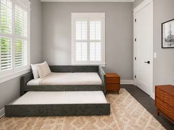 Sunlit residential bedroom with gray upholstered daybed and pulled-out trundle, striped pillow, wooden nightstands, white plantation shutters, beige shag rug and light gray walls.