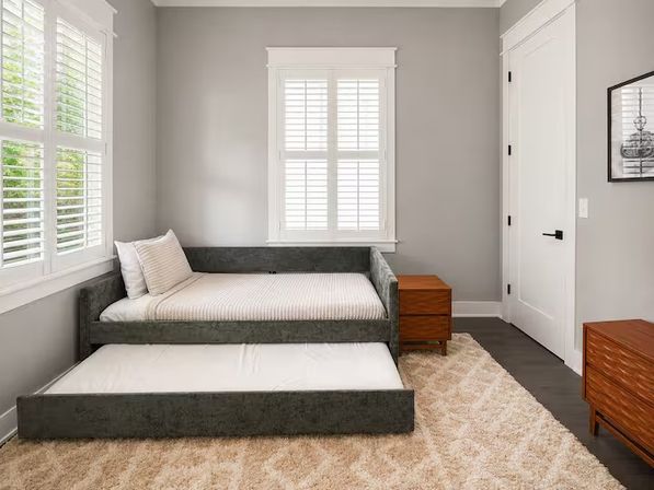 Sunlit residential bedroom with gray upholstered daybed and pulled-out trundle, striped pillow, wooden nightstands, white plantation shutters, beige shag rug and light gray walls.