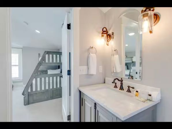 Bright modern home interior showing a white marble bathroom vanity with bronze faucets and wall sconces, mirror, and an open door revealing a bedroom with gray wooden bunk beds.