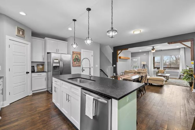 Open-concept modern kitchen with large black island, stainless steel appliances, white cabinets, pendant lights and hardwood floors opening to a bright living room with a sectional and large windows.