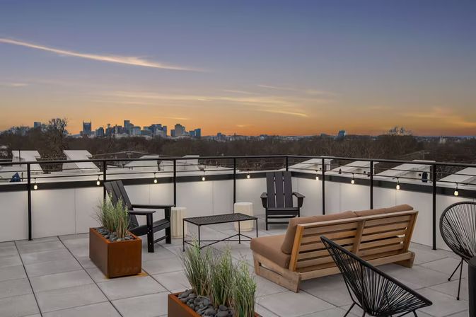 Cozy rooftop terrace with modern lounge chairs, wooden bench, planters and string lights overlooking a distant city skyline at sunset