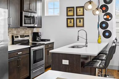 Modern kitchen with dark wood cabinets and stainless steel appliances, white quartz breakfast bar with undermount sink and flexible-neck faucet, black leather bar stools, pendant lights, tiled backsplash, coffee maker and vinyl record wall decor.